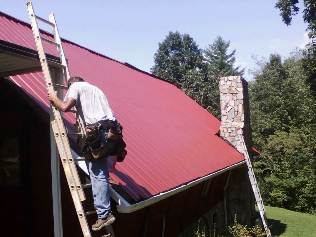 Roofer on a ladder beside a red metal roof and stone chimney — Soffit & Fascia Repair by Roofing Brothers in Hubbard